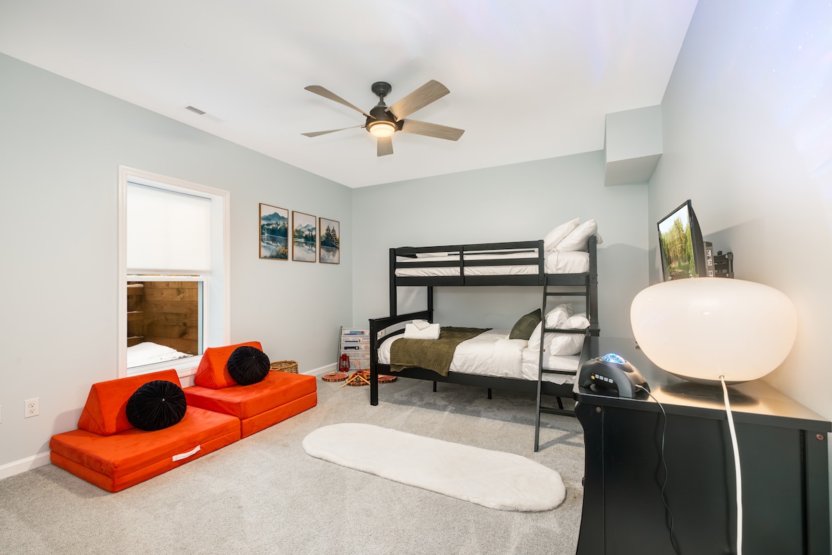 Svijetla dječja soba s krevetima na kat, dva crvena madraca za izgradnju tvrđave ili dodatni prostor za spavanje, projektor sa zvijezdama, dječje knjige, mekani tepih, umjetnost s planinama i fenjer s lažnom vatrom za izmišljene priče. Savršeno za igru i ugodne filmske večeri.