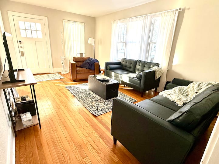 วิวจากทางเดินด้านหลังห้องนอน วิวประตูหน้าบ้านไปยังระเบียง โซฟาสองตัวและเก้าอี้นวมหนึ่งตัว ตะกร้าเกม ทีวีที่เชื่อมต่อ Wi-Fi ลำโพงบลูทูธ ที่นอนขนาดเต็มในหีบสีดำ เตียงสุนัขสองเตียง ที่แขวนเสื้อโค้ท และร่ม 