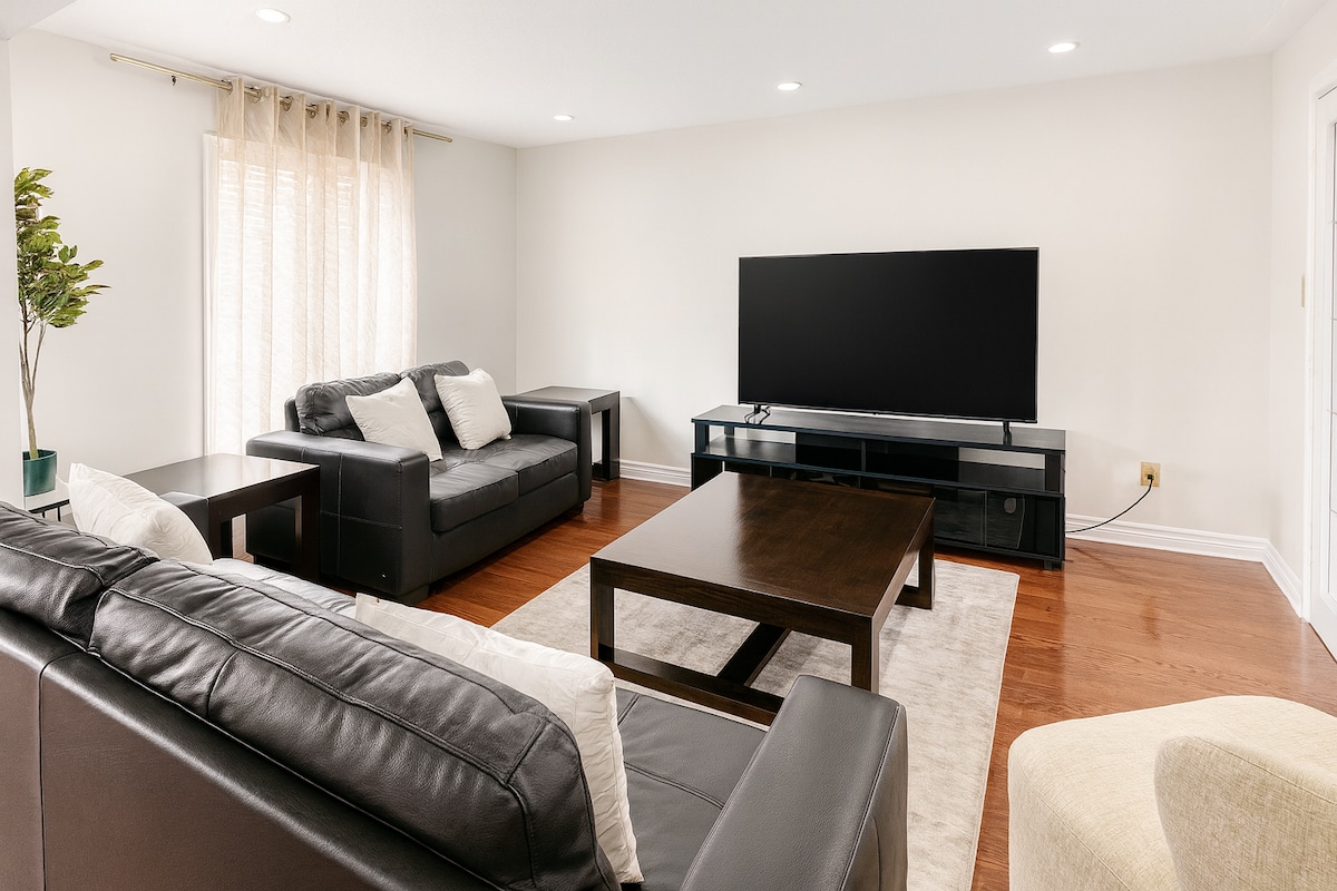 غرفة معيشة عصرية مع أرائك جلدية سوداء أنيقة وتلفزيون بشاشة مسطحة كبيرة وأرضيات خشبية دافئة. تخلق الإضاءة المريحة والديكور البسيط مساحة مريحة مثالية للاسترخاء أو مشاهدة الأفلام.