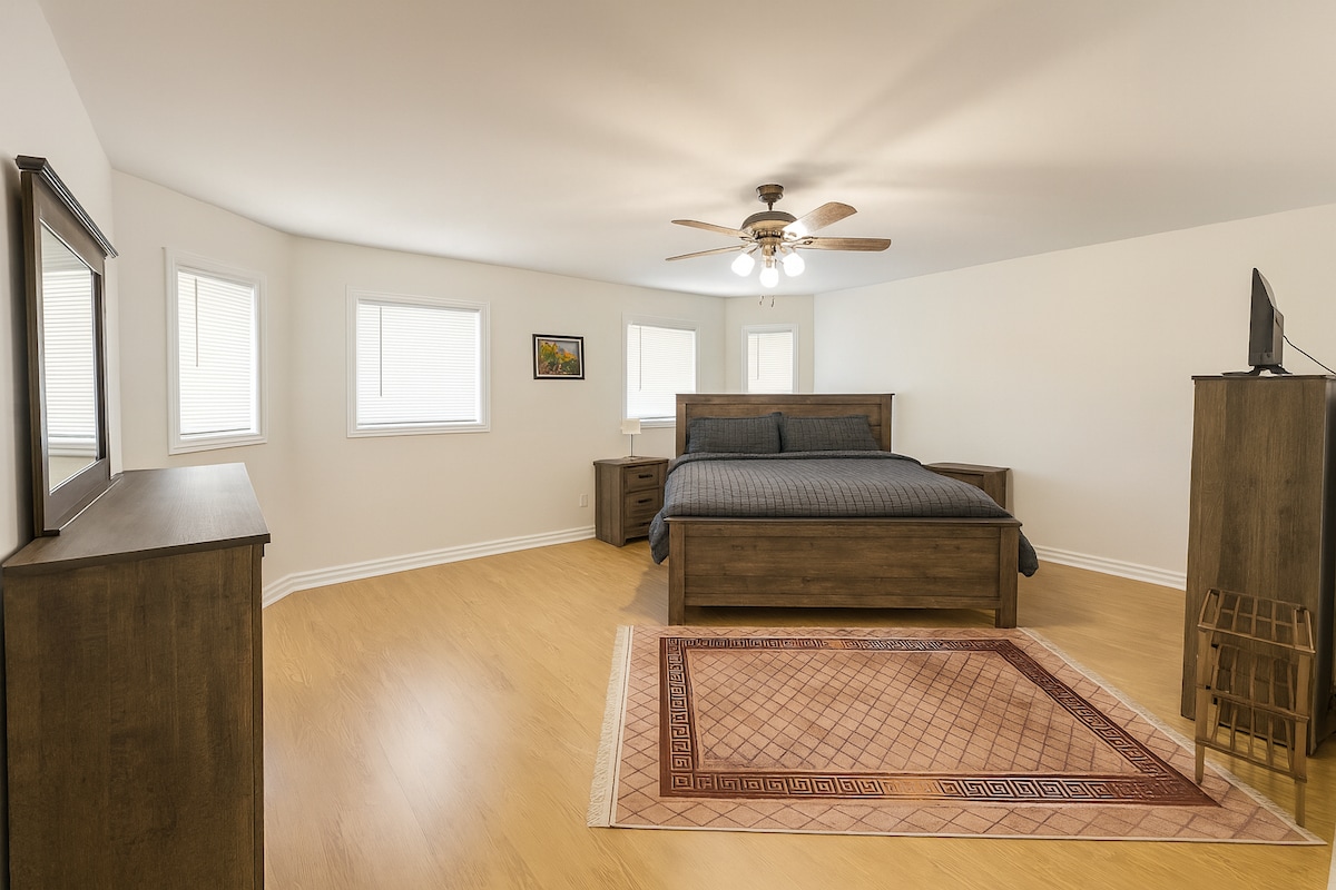 غرفة نوم رئيسية واسعة تتميز بسرير كينج ومروحة سقف وخزانة ملابس مع مرآة وتلفزيون. تملأ النوافذ الكبيرة الغرفة بالضوء الطبيعي، مما يخلق ملاذًا دافئًا ومريحًا للضيوف.