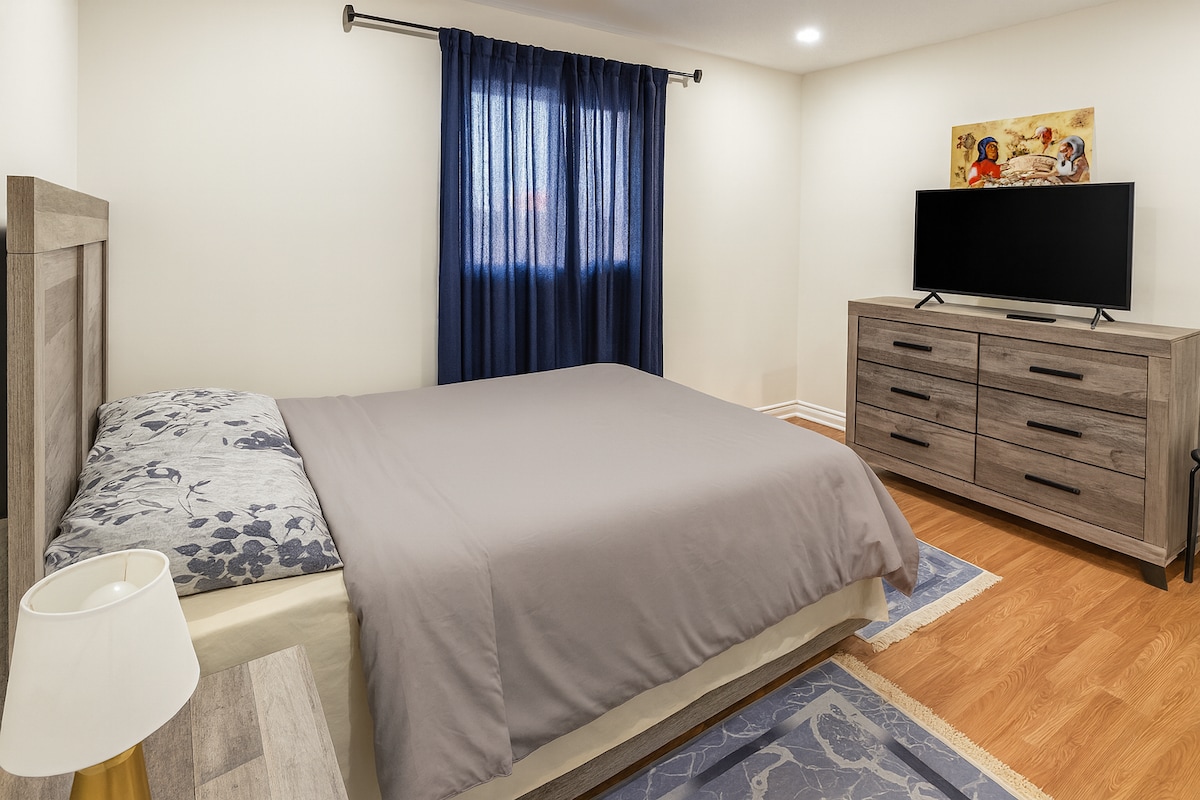 غرفة نوم أنيقة مع سرير مريح بحجم كبير، وأثاث خشبي رمادي أنيق، وسجاد ناعم، وتلفزيون بشاشة مسطحة - مثالية لقضاء ليالٍ مريحة وصباح مريح.