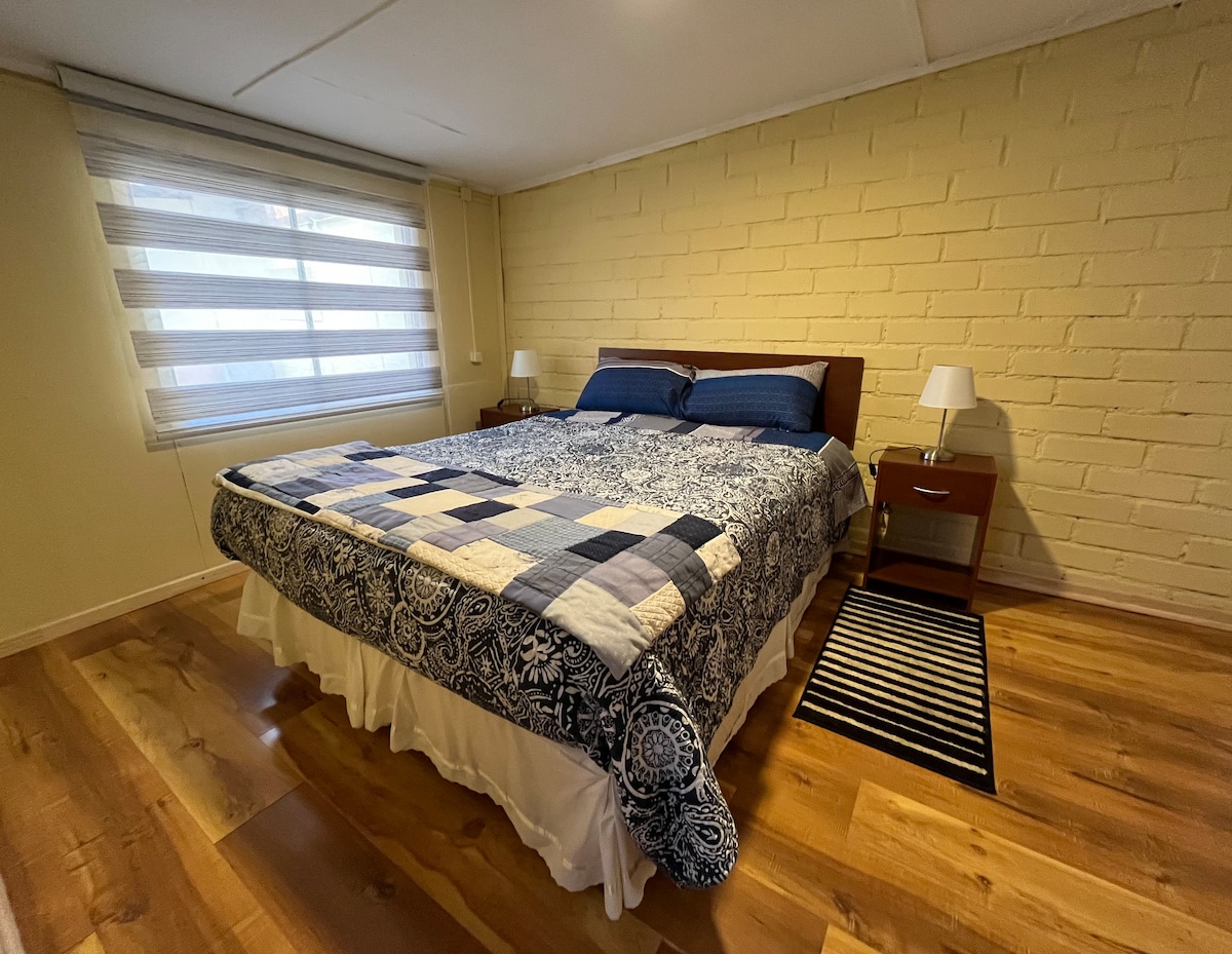 חדר גדול עם מיטה זוגית עם שידות לילה, מנורות, שטיח ליד המיטה ומתלה בגדים.