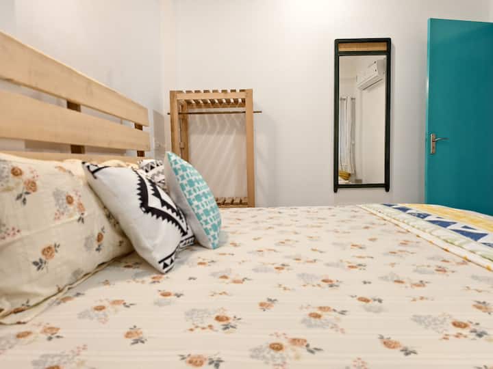 Chambre spacieuse décorée esthétiquement avec un lit confortable et un miroir sur le mur. Un support de rangement est également là. Plus grande fenêtre dans la chambre avec vue sur Mandir