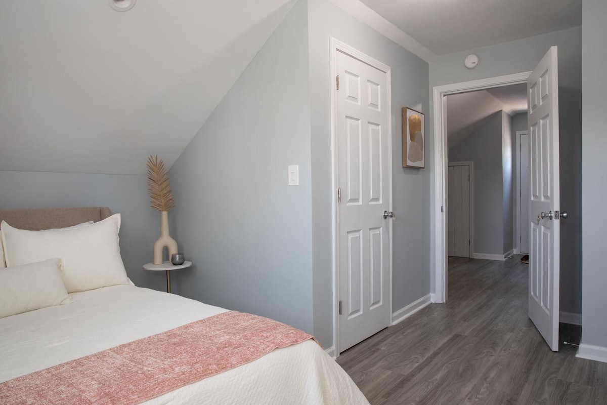 Pokojná spálňa v podkroví s posteľou s bielou a ružovou posteľnou bielizňou a dekoratívnou vázou na malom bočnom stolíku. Svetlomodré steny a biele dvere vytvárajú pokojnú atmosféru s náhľadom do ďalšej miestnosti na chodbe.