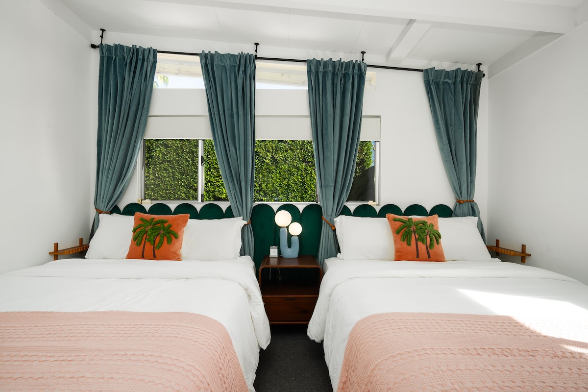 Ova elegantna spavaća soba ima dva bračna kreveta s elegantnim valovitim uzglavljem i nježnim detaljima u boji ruža.