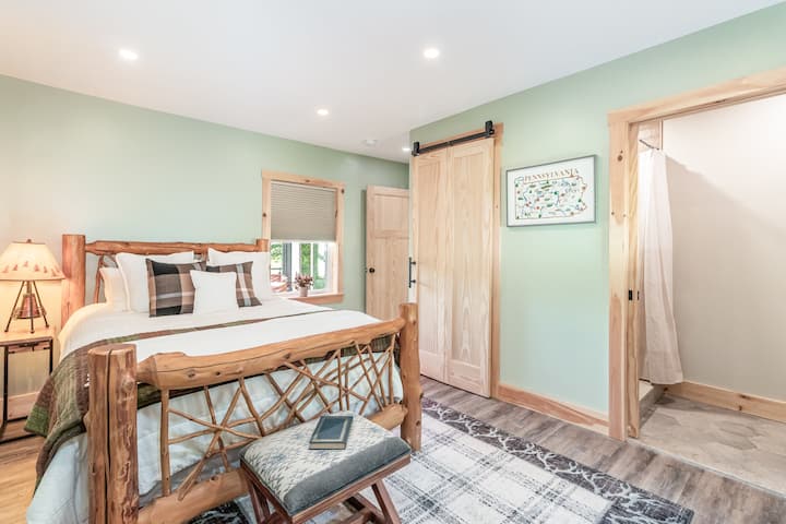 Cozy second bedroom featuring a rustic queen log bed, soft layered bedding, and calming sage-green walls. Includes a sliding barn door, plush area rug, bedside lighting, and private access to the nearby bathroom for added convenience.