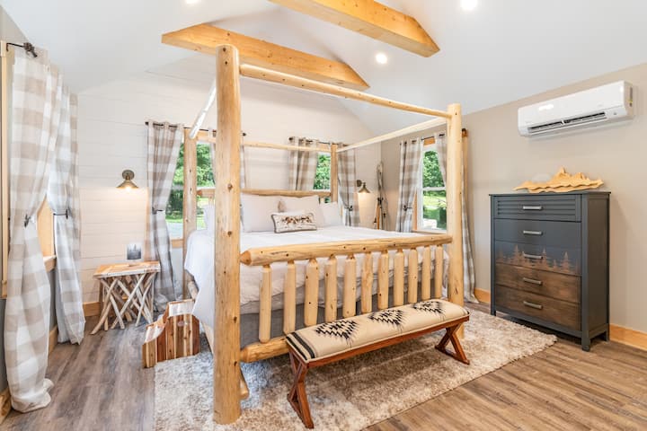 Rustic king bedroom with a handcrafted log canopy bed, vaulted ceilings, and exposed wood beams. Soft lighting, cozy rug, and large windows with scenic views create a peaceful retreat. Includes climate control and stylish dresser for storage.