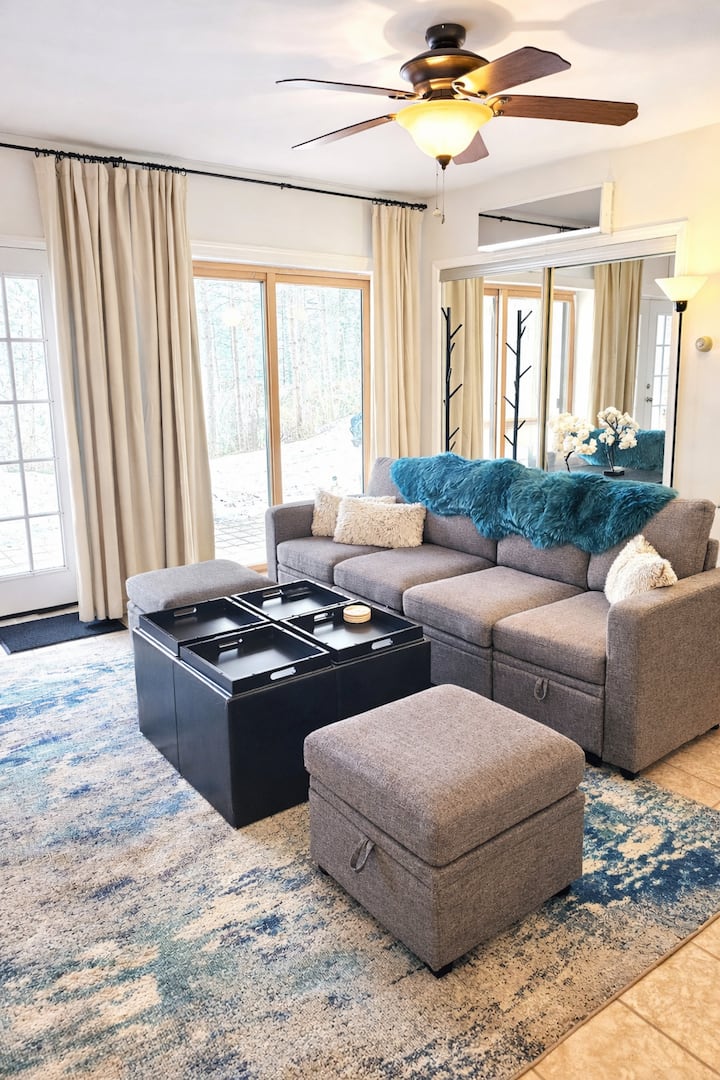 It features a comfy queen sleeper sofa and extra ottomans for the whole family. Enjoy movie nights in this sun-lit living room! Large windows offer serene lake views, creating the perfect spot to relax and recharge. Ideal for your next getaway!
