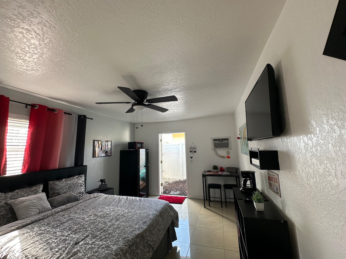 A private room features a spacious bed with neutral bedding and a decorative throw. A ceiling fan hangs above, and large windows are dressed with red curtains. A kitchenette is visible, along with a wall-mounted television, and the entrance to the room opens to an outdoor space.
