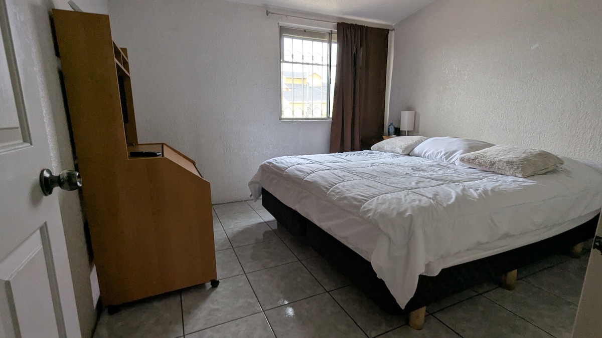 A spacious bedroom features a large bed with crisp white bedding and two pillows. Natural light filters through a nearby window, accented by dark curtains. A wooden television stand sits against the wall, adding functionality to the room's simple decor.