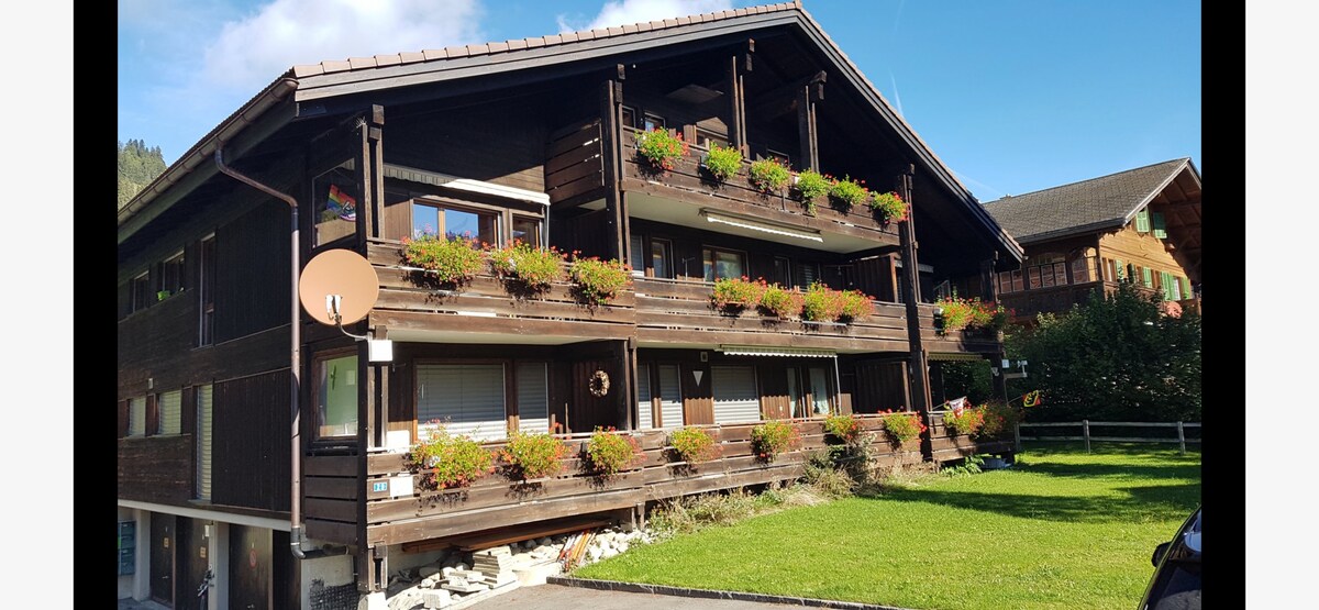 A multi-family building is shown, featuring multiple balconies adorned with vibrant flowers. Lush green grass surrounds the structure, and several windows allow natural light to enter. The architecture is traditional, with wooden paneling and a sloping roof that enhances the overall aesthetic.