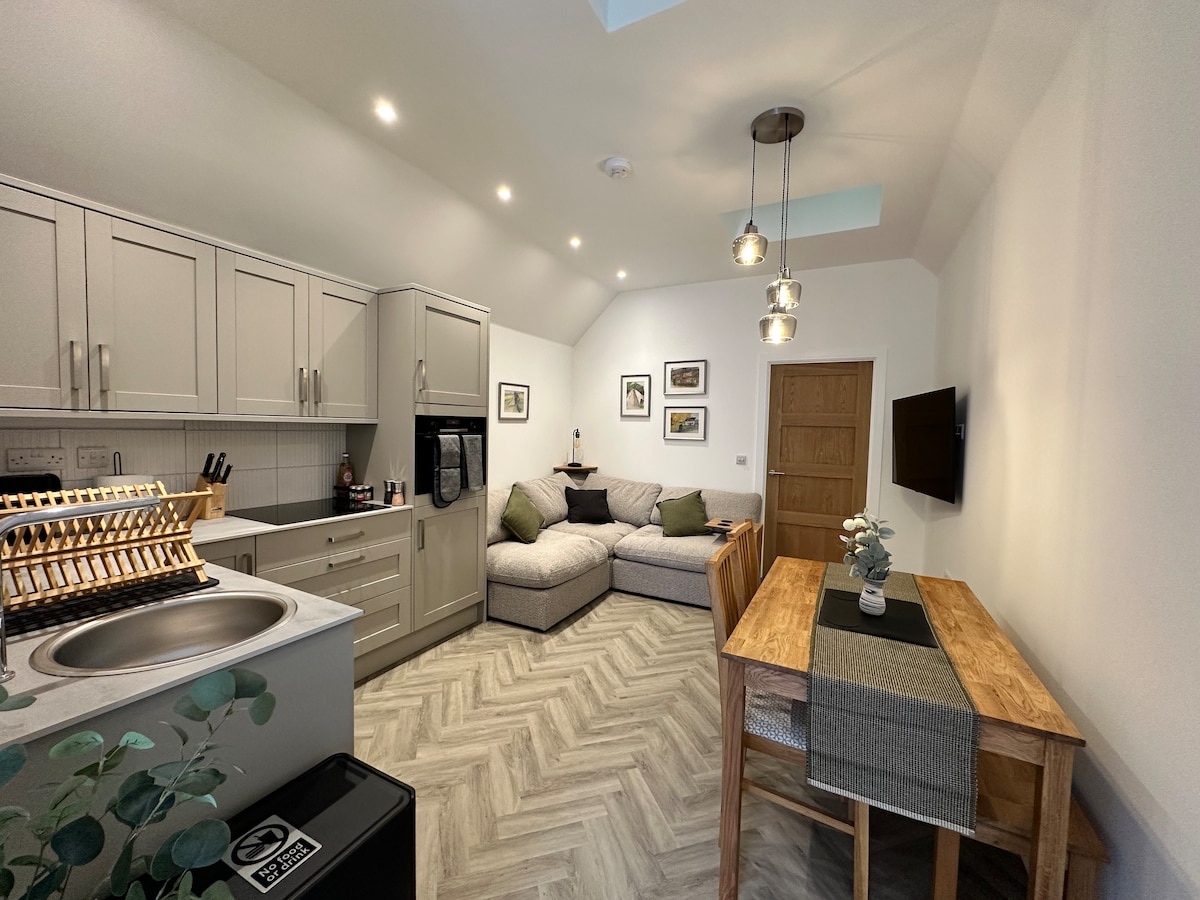 A newly built open-plan living area is displayed, featuring a modern kitchen with light cabinetry and a washing machine. A spacious sofa is positioned in front of a wall-mounted TV, while a wooden dining table with seating stands nearby. Natural light floods the space through skylights.