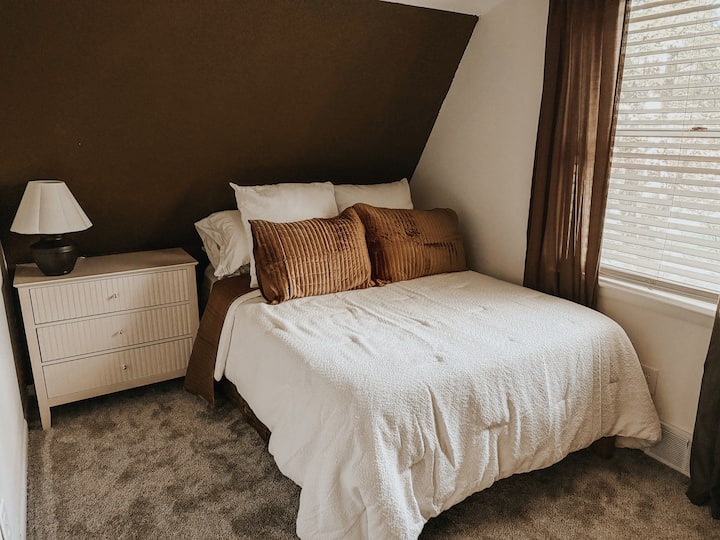 Útulná spálňa na poschodí so šikmým stropom v štýle chaty, mäkkou posteľnou bielizňou a teplými neutrálnymi tónmi – ideálna na pokojnú noc.