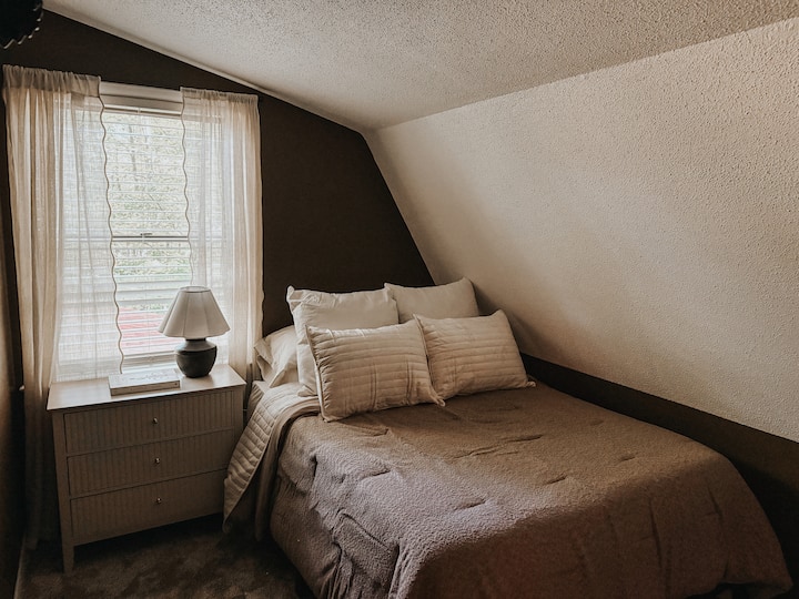 Teplá spálňa v štýle horskej chaty na poschodí s viacvrstvovou posteľnou bielizňou, mäkkým osvetlením a pokojným výhľadom z okna – ideálna na pokojné rána.