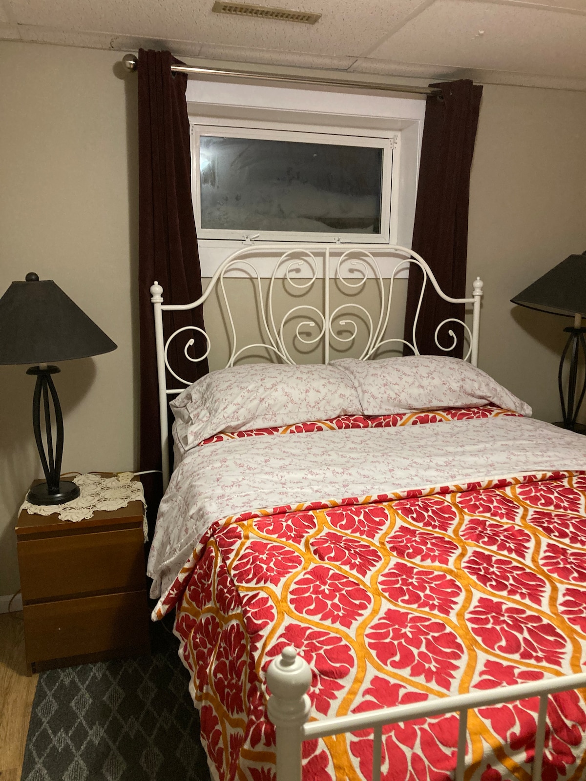 A cozy sleeping area features a white metal bed framed by decorative details. A patterned red and orange blanket covers the spacious mattress. Two bedside lamps provide soft lighting, and a small wooden nightstand rests beside the bed. A window with dark curtains allows natural light to enter.