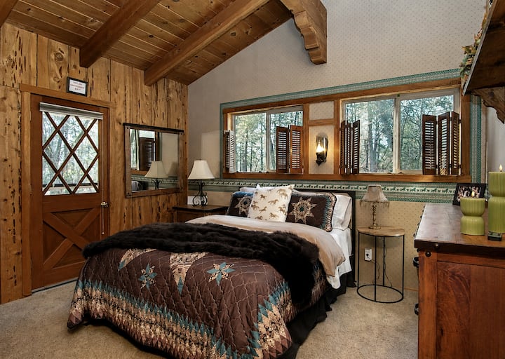Unwind in The Bridle Room's rustic elegance. This cozy, wood-paneled sanctuary features a plush queen bed under vaulted exposed beams, classic Western decor, and custom window shutters. With a private exterior door, it’s the ultimate retreat.