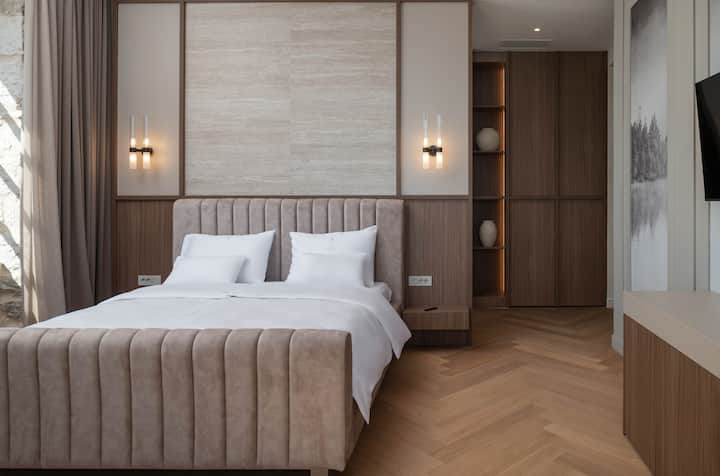Првата спална соба со брачен кревет (широк 180-220 см), рачно изработен дизајнерски мебел и големи прозорци со мирни погледи, комбинирајќи удобност, елеганција и безвременски медитерански стил за одморно прибежиште.