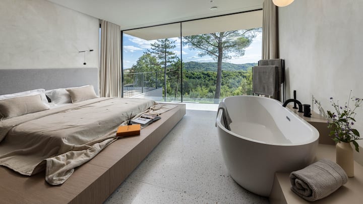 Dormitorio principal de lujo en Villa L Excelsior con cama tamaño king, baño privado y baño independiente junto a grandes ventanales. Despierta con impresionantes vistas a los viñedos y al bosque, combinando elegancia, comodidad y naturaleza serena desde tu habitación.
