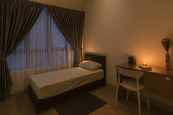 Dormitorio luminoso y acogedor con grandes ventanales, elegantes cortinas grises y suave luz natural. Cuenta con una cómoda cama individual, escritorio de madera con silla, jarrón con flores e iluminación cálida, perfecta para relajarse o trabajar en un espacio moderno y tranquilo.