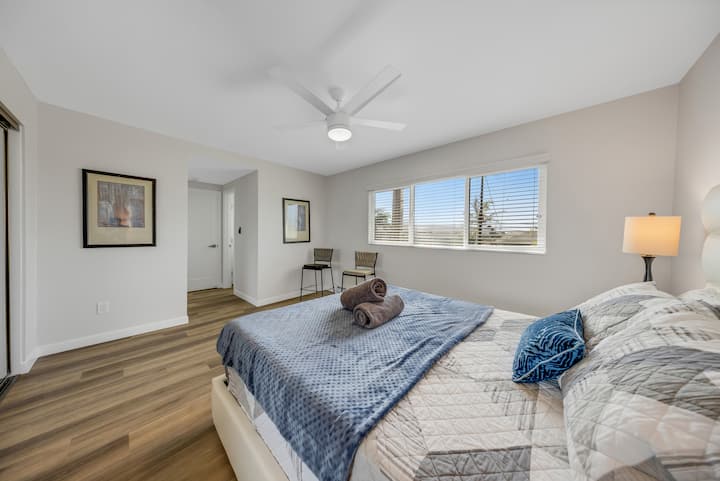 Një dhomë gjumi e rehatshme me një krevat të rregulluar mirë me shtroja blu dhe gri, një tavolinë të vogël me një karrige pranë dritares dhe një abazhur në tavolinën anësore. Dhoma ka dysheme druri, një ventilator tavani dhe vepra arti të kornizuara në mur, duke krijuar një atmosferë të qetë.