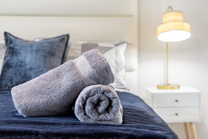 Две уредно свиткани сиви крпи се потпираат на плишана прекривка за кревет, со стилска ноќна ламба и маса во заднина, создавајќи удобен амбиент во спалната соба.