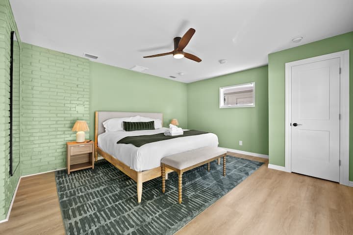 Štýlová izba s pohodlnou manželskou posteľou, šalviovo zelenými stenami a textúrovaným tehlovým akcentom. Užite si prirodzené svetlo, mäkký koberec a moderný stropný ventilátor pre útulný a relaxačný pobyt.