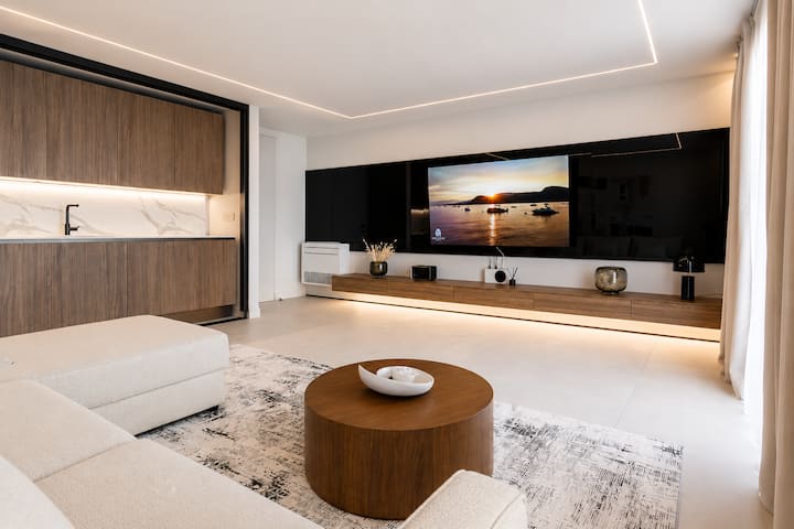 Golfo Aranci'deki AZULIS Athos Villa'da modern oturma odası; bej renkli çekyat (2 kişilik), yuvarlak ahşap sehpa, X-Box oyun konsollu TV duvarı, küçük mutfak, perdeler, ekstra giriş ve LED tavan aydınlatması.