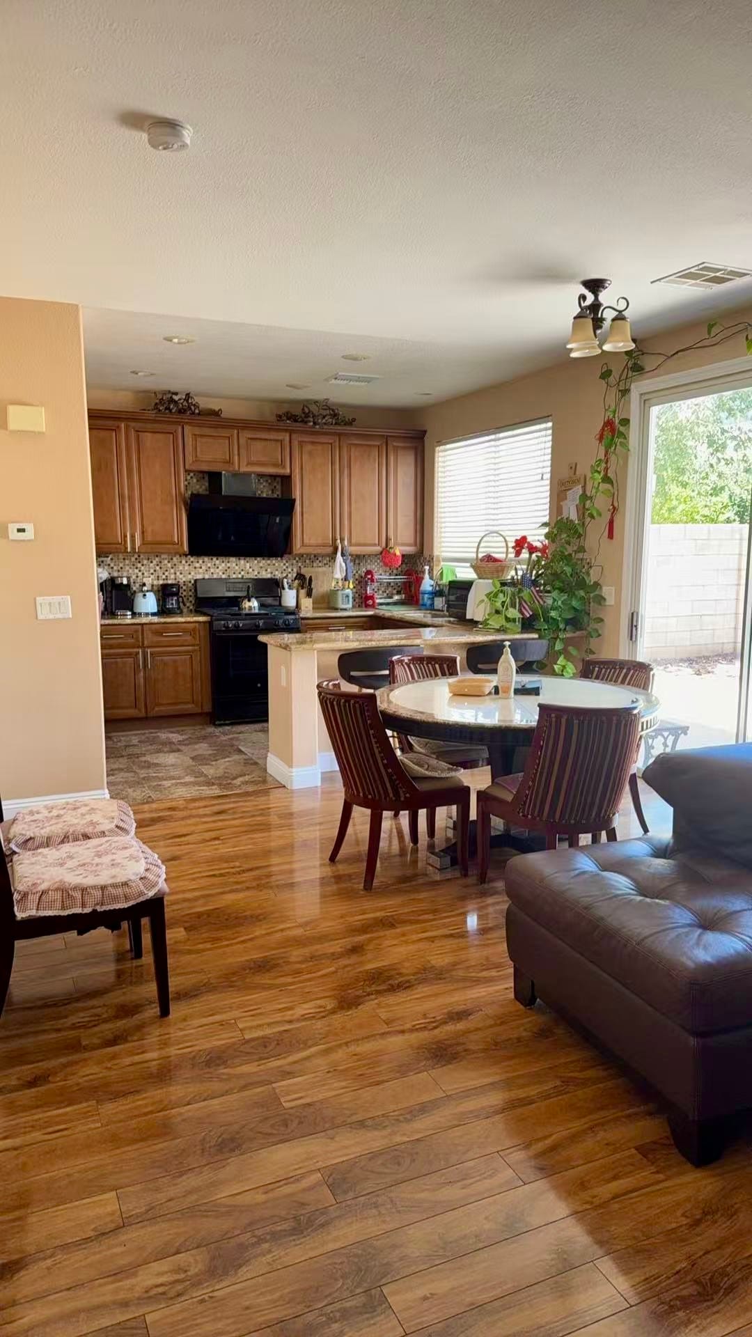 A modern kitchen area is visible, featuring dark wooden cabinetry and a full set of appliances. A round dining table with dark chairs accommodates several people. Natural light enters through large glass doors, enhancing the wood laminate flooring and creating a welcoming space.