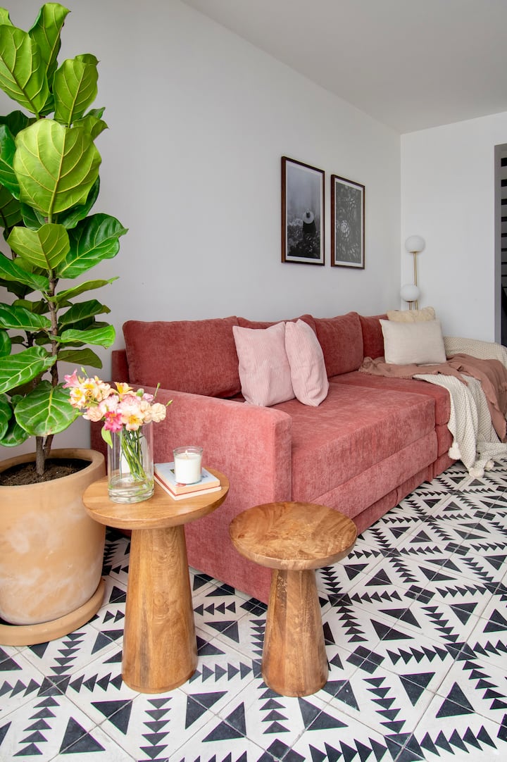 Მისაღები ოთახის დახვეწილი კუთხე, რომელშიც არის რბილი ტერაკოტის დივანი, რომელიც ჩარჩოშია მოქცეული გეომეტრიული იატაკითა და ხის ბუნებრივი აქცენტებით. Აყვავებული სიმწვანე და დახვეწილი დეკორი ქმნის მყუდრო, მაგრამ დიზაინერულ ატმოსფეროს, რომელიც უპრობლემოდ კურირებს.