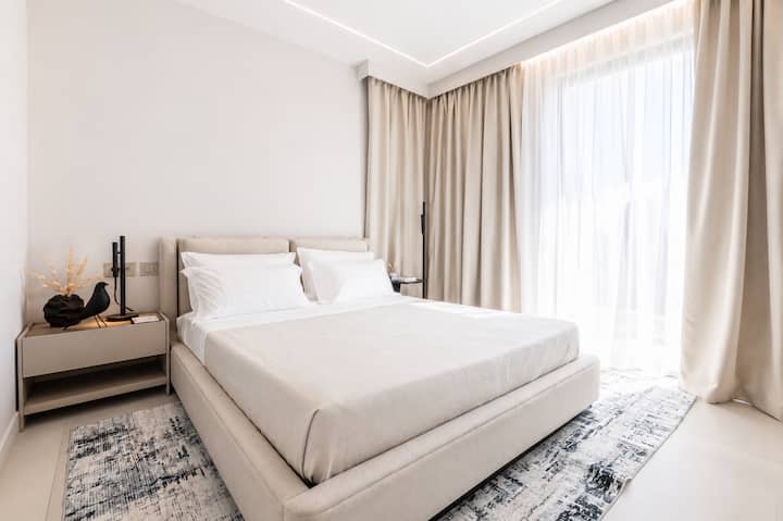 Golfo Aranci'deki AZULIS Aramis Villa'da büyük döşemeli yatak, nötr renkli yatak takımı, komodin, yumuşak perdeler, halı ve doğal ışık bulunan aydınlık ve modern bir yatak odası.