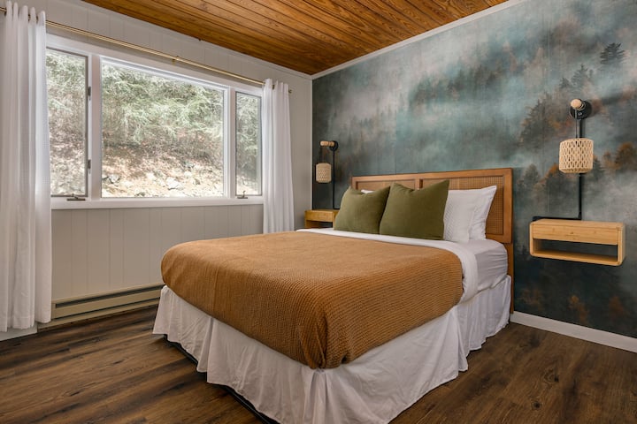 חדר שינה עם מיטת קווין, מצעים נוחים מכותנה ונוף שקט ליער דרך חלונות גדולים.