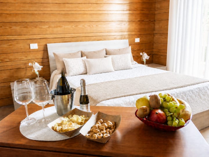 Camera matrimoniale calda e romantica, con pareti in legno e letto dai toni panna. Calici, bollicine e piccoli assaggi trasformano la stanza in un invito a rallentare e godersi un aperitivo da condividere in due, in un’atmosfera intima e rilassata.