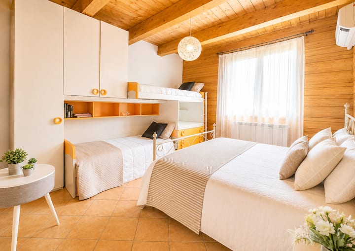 Camera ampia e luminosa, dove il legno incontra i toni chiari degli arredi. Letto matrimoniale e due letti singoli offrono spazi confortevoli, mentre la grande finestra e le tende bianche lasciano entrare una piacevole luce naturale.
