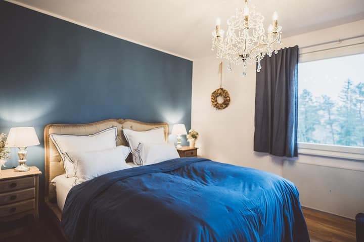 ห้องนอนหรูหราพร้อมเตียงคู่ที่นุ่มนวล โต๊ะข้างเตียงที่มีสไตล์ และโคมระย้า ผนังสีฟ้าสดชื่น หน้าต่างที่มีวิวธรรมชาติ และการตกแต่งอย่างมีรสนิยมให้บรรยากาศที่ผ่อนคลายและความสะดวกสบายอย่างเงียบสงบ