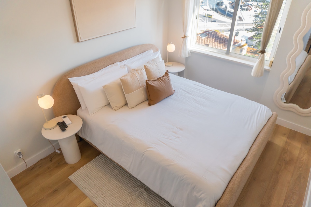 მსხვილი კადრი, რომელზეც ჩანს ნათელი, ელეგანტური მთავარი საძინებელი, კომფორტული საწოლი, თანამედროვე დეკორი და ფანჯრიდან ხედი.