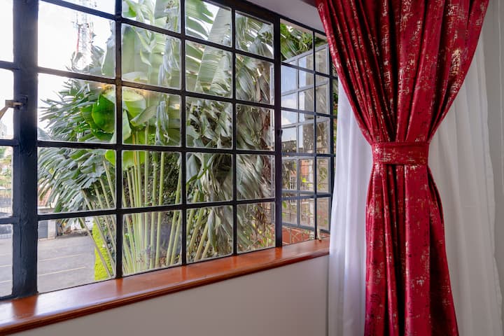 Vista del dormitorio junto a la ventana con vista a la vegetación, enmarcada con cortinas rojas que agregan calidez y privacidad a la habitación.