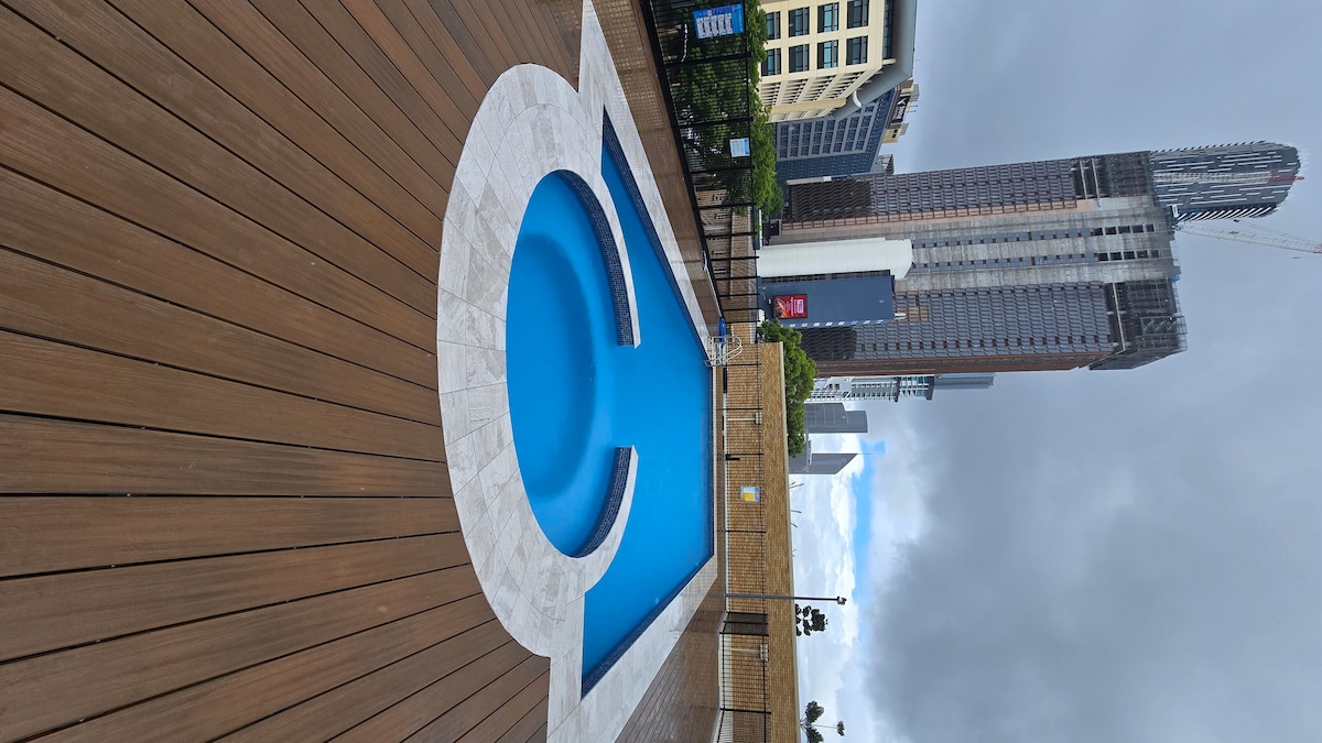 A swimming pool is surrounded by wooden decking, featuring a unique circular design within its rectangular shape. The clear blue water contrasts with the beige stone border. Tall buildings rise in the background, providing an urban backdrop to the pool area.