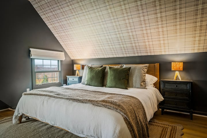 Cozy up under the eaves: This unique guest suite features a plush king bed nestled beneath a striking plaid vaulted ceiling—the ultimate den for a restful night’s sleep. ✨