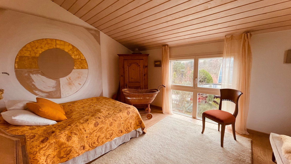 A spacious room is featured, with a large bed dressed in a warm, patterned coverlet. A wooden cupboard provides storage, and a vintage chair sits nearby. Large windows are adorned with light curtains, allowing natural light to fill the room while a cozy rug adds comfort underfoot.