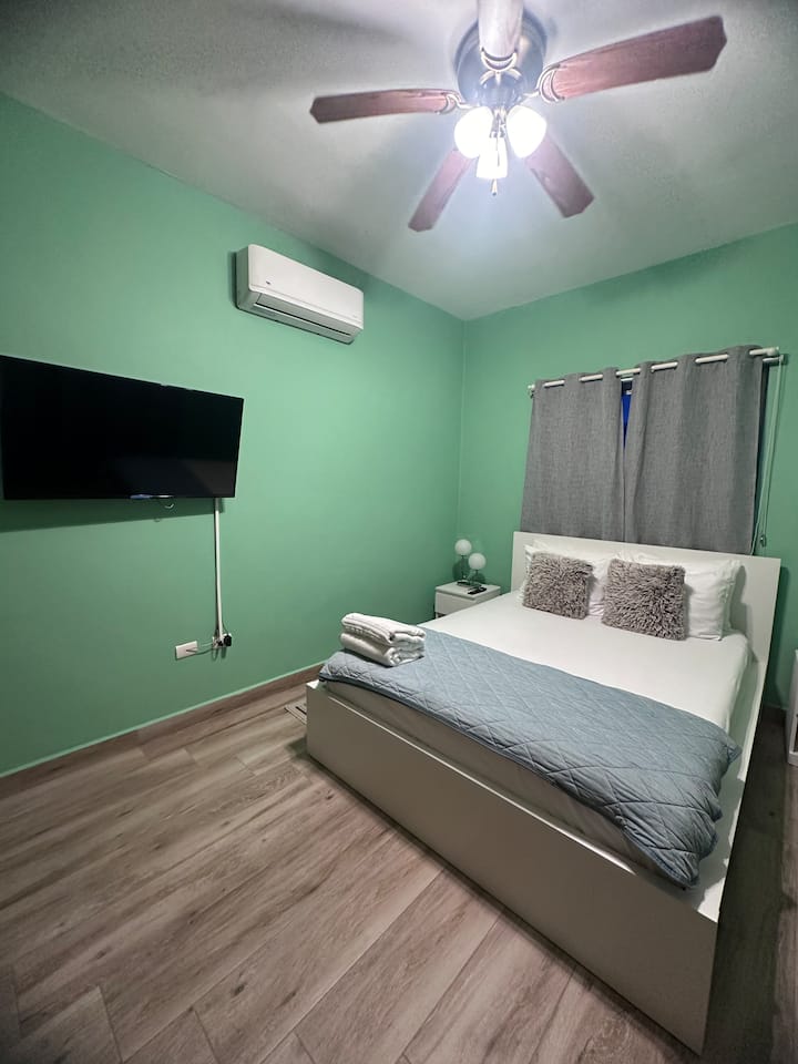 Comfortable bedroom with a queen bed, air conditioning, and a wall mounted TV. Soft lighting and simple furnishings create a quiet, practical space for a restful stay.
