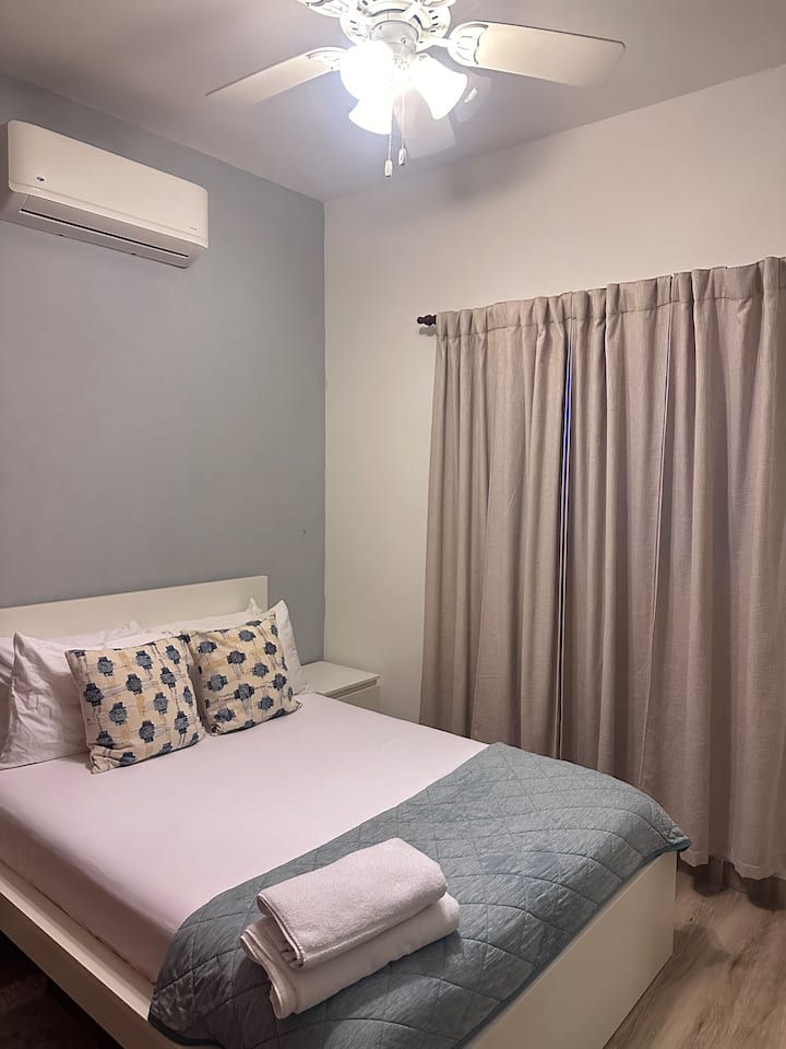 Cozy bedroom with a comfortable bed, air conditioning, and blackout curtains for a restful sleep. Soft lighting and a simple layout create a calm and private space to unwind.
