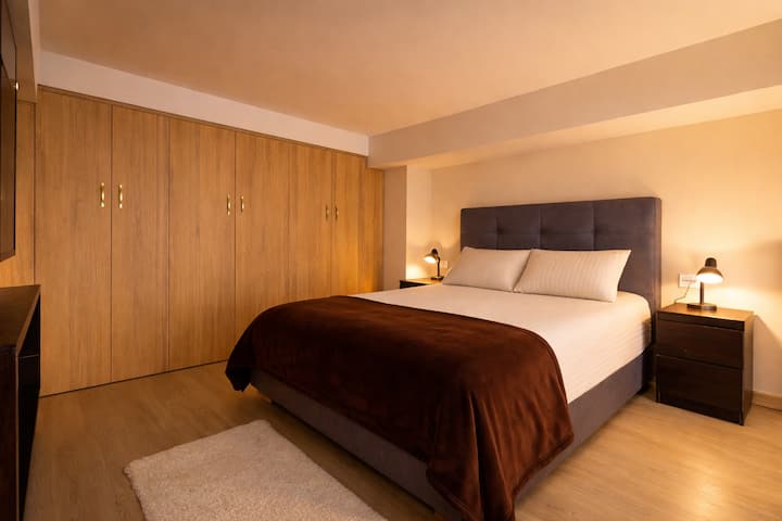 Gezellige kamer met een tweepersoonsbed, warme verlichting en een grote houten kast. De eenvoudige inrichting en de neutrale kleuren zorgen voor een ontspannen en elegante sfeer, ideaal om uit te rusten en te genieten van een comfortabel en vredig verblijf.