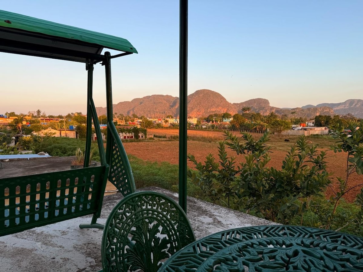 A serene terrace is shown with a green swing and a round table surrounded by intricately designed chairs. A scenic view of lush fields and distant mountains is visible in the background, with soft light illuminating the landscape as dusk approaches.