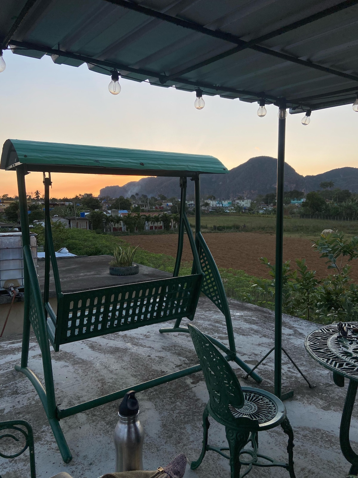 A swing and two comfortable chairs are positioned on a rooftop terrace. The background features a scenic view of mountains and farmland, with soft sunset hues in the sky. Decorative string lights add a cozy touch to the setting.