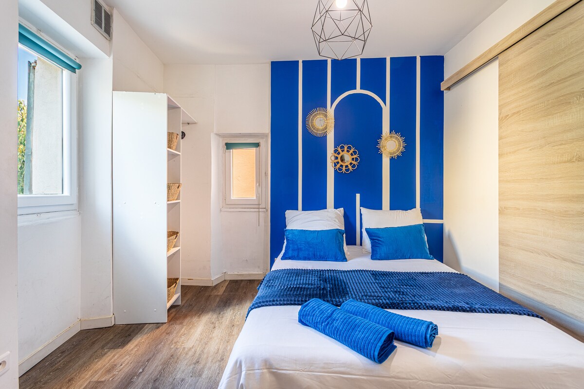 Pogled s desne strane na bračni krevet i plavi zid s bijelim ukrasnim letvicama. Drveni pod, veliki prozori i minimalistički dekor sa zlatnim i plavim detaljima koji podsjećaju na mediteranski duh.