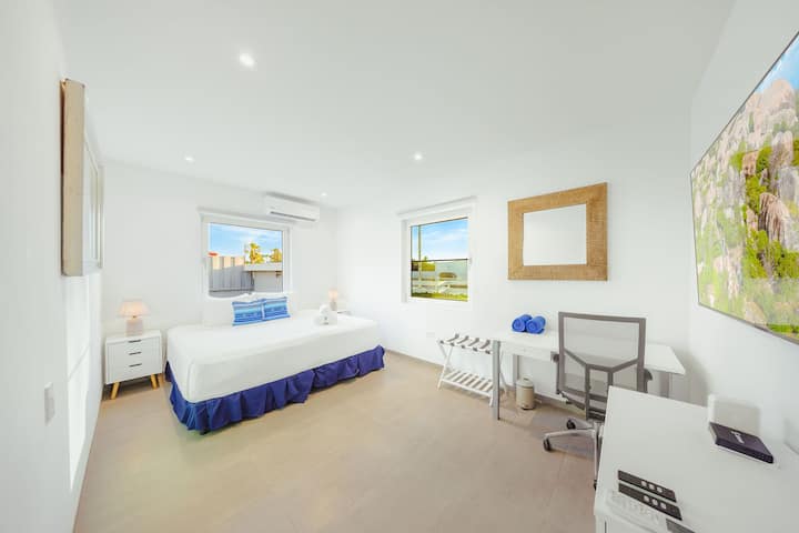 Quarto iluminado com ar condicionado, smart TV, cômoda e espaço de trabalho — perfeito para conforto e trabalho remoto.
