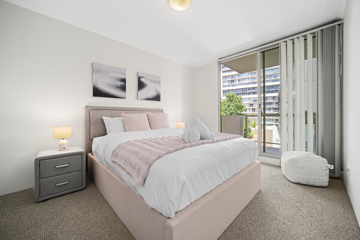 غرفة نوم مشرقة وأنيقة بألوان ناعمة وأغطية فاخرة وإمكانية الوصول إلى الشرفة. استيقظ على الضوء الطبيعي والإطلالة الحضرية الهادئة.