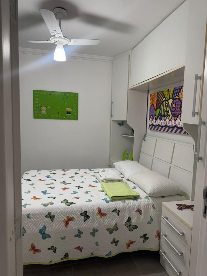 Spálňa s manželskou posteľou, šatníkom, zrkadlom, vankúšmi, posteľnou bielizňou a prikrývkami má okno so žalúziami a stropným ventilátorom.   