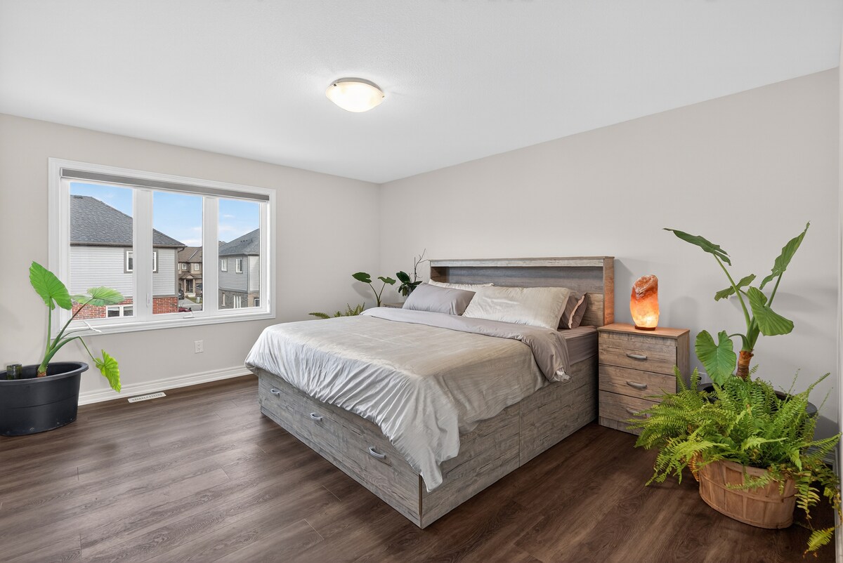 Пространа спална соба со големи прозорци, удобен брачен кревет (широк 180-220 см) со вградени фиоки за складирање и мека неутрална постелнина. Собните растенија и топлата ламба покрај креветот додаваат нежен, релаксирачки допир на светлиот, отворен простор.