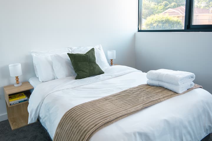 Cómoda cama doble con sábanas blancas y suaves, almohadas mullidas y una manta con textura. Las mesitas de noche con lámparas suaves crean un espacio cálido y acogedor, mientras que la luz natural fluye desde la gran ventana de arriba.